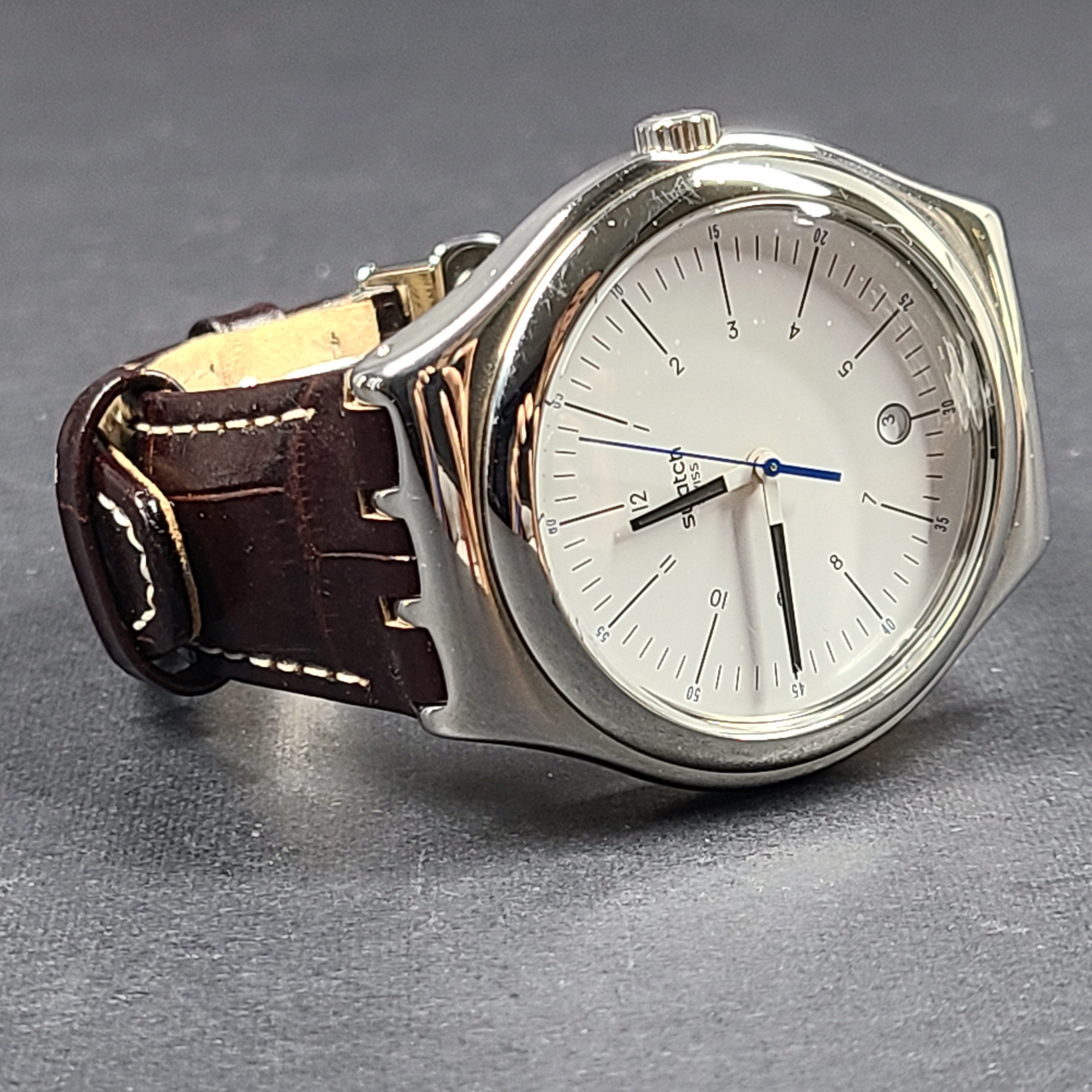 Swatch YGS452G stainless steel wristwatch with a white dial and brown leather strap.