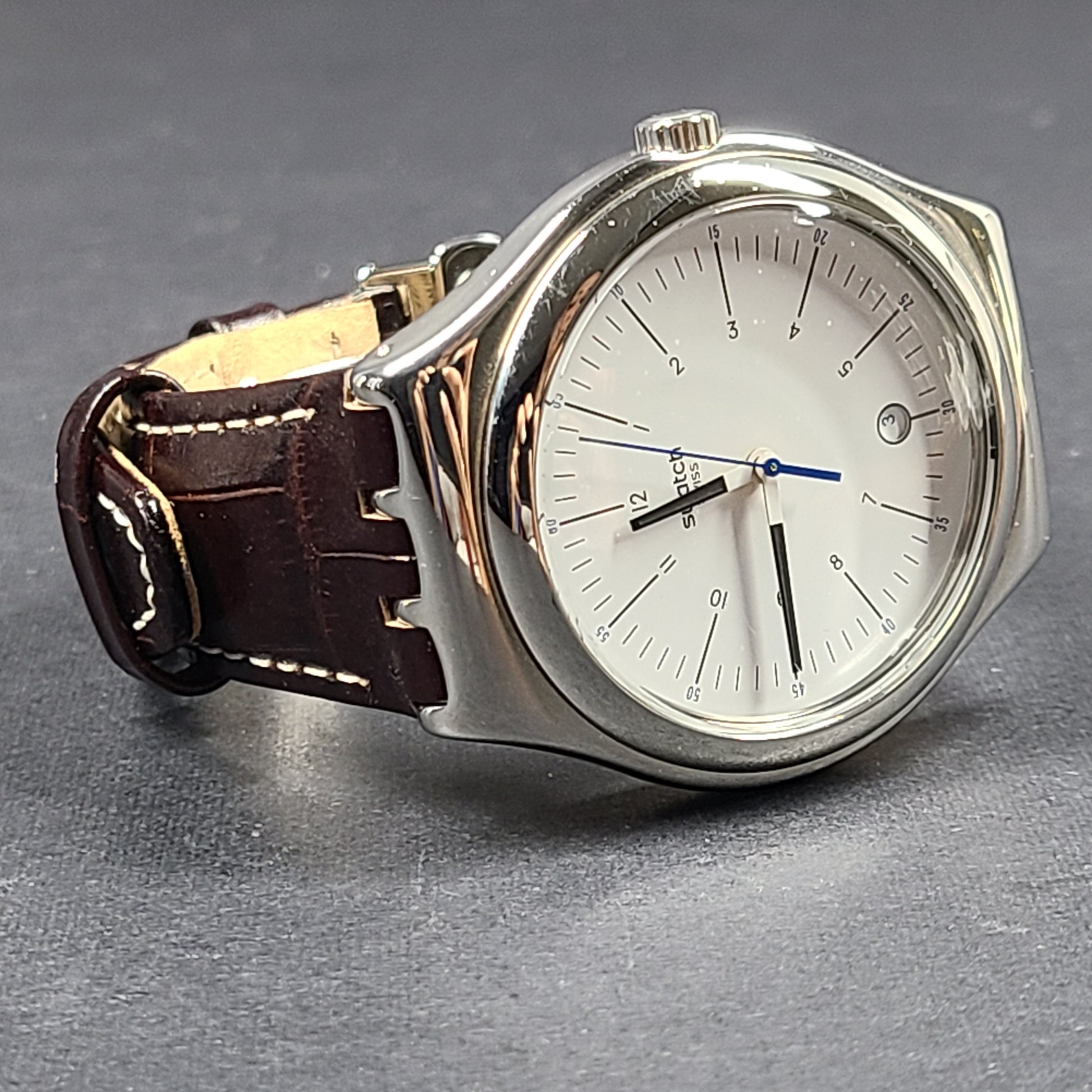 Swatch YGS452G stainless steel wristwatch with a white dial and brown leather strap.